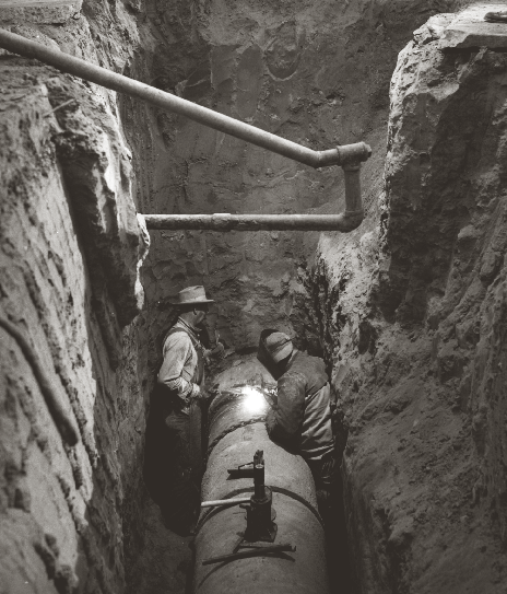 Historical black-and-white photograph of two workers welding a large water main pipeline inside a deep earthen trench, circa early 20th century