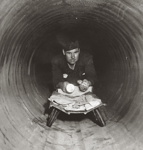 Historical black-and-white photograph of a Cal Water employee inside a large water main, reading blueprints by flashlight